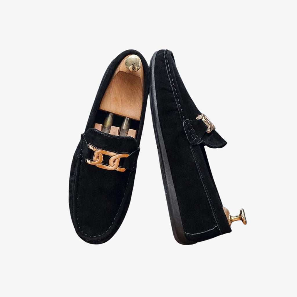Zwarte herenloafers van synthetisch suède, elegant en modern, met een gouden metalen gesp op de voorkant. Het verfijnde design toont zichtbare stiksels die karakter toevoegen. De zwarte zool maakt de sobere en chique uitstraling compleet.