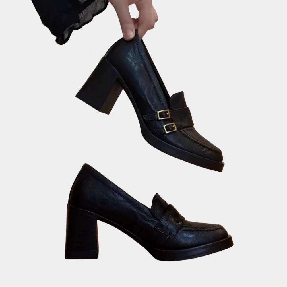Paar zwarte loafers met hoge hak, waarvan een schoen van opzij staat en de tweede tussen de vingers van een vrouw wordt vastgehouden