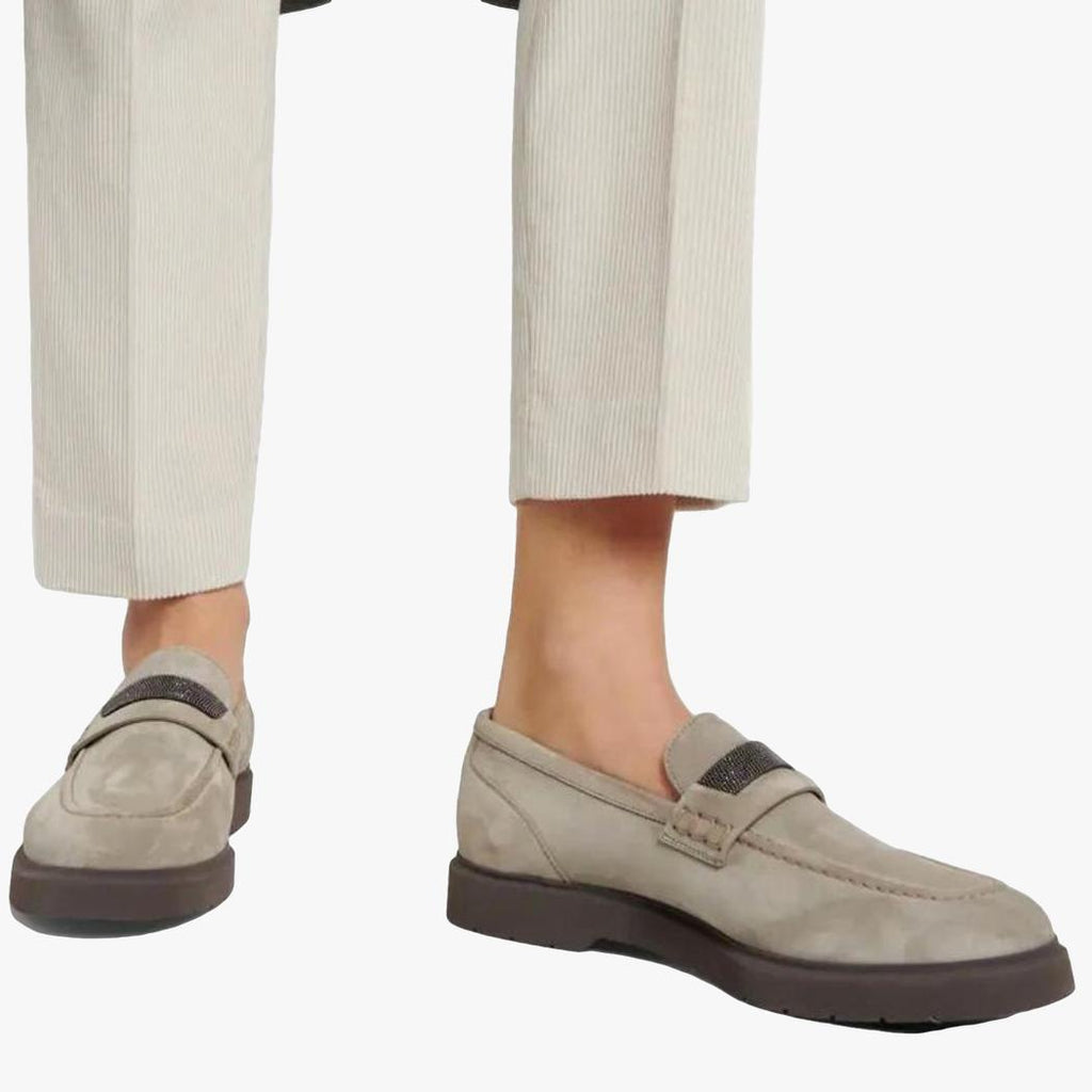je ziet de voeten van een vrouw met een beige pantalon en grijze dames loafers van echt suède