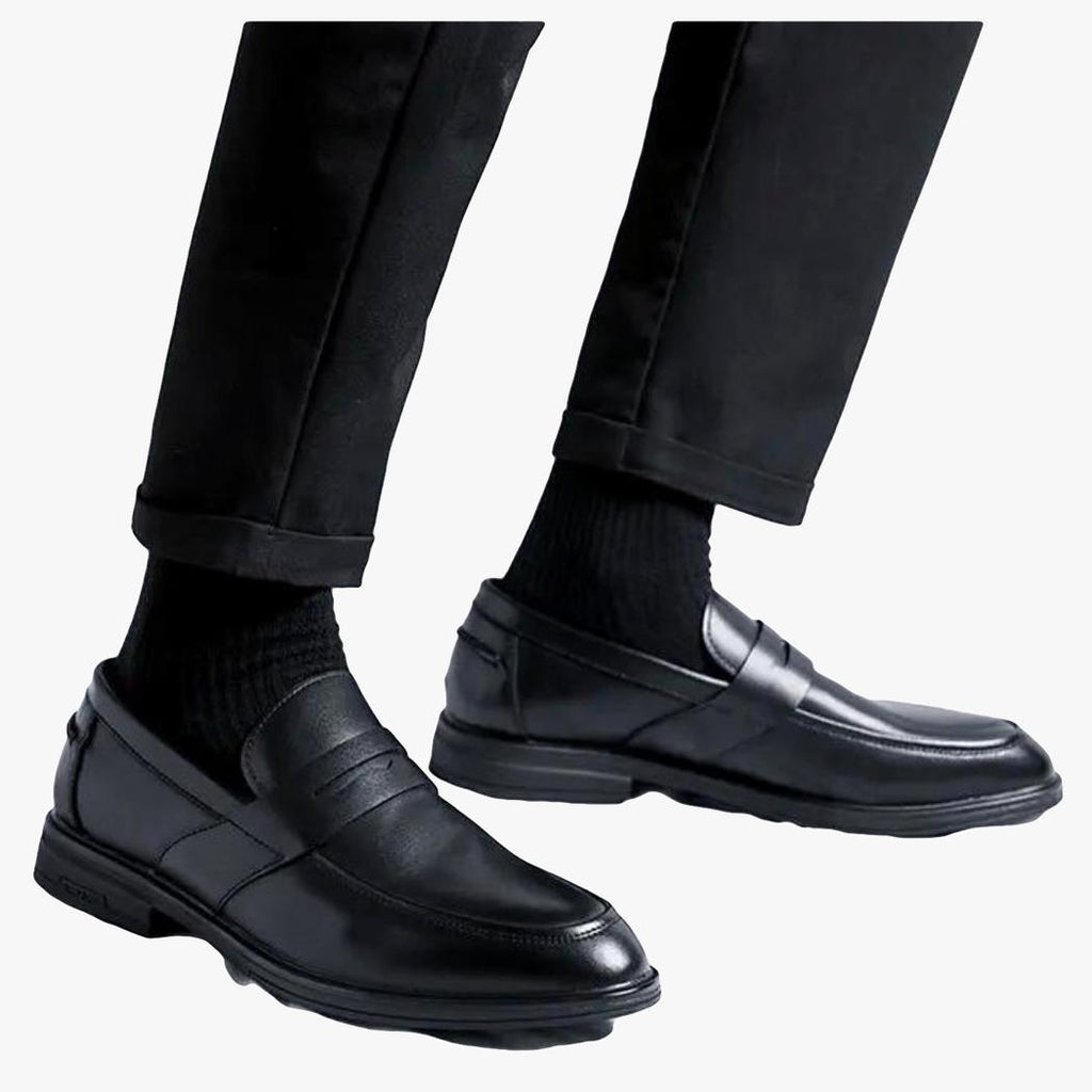 je ziet op een lichte achtergrond de onderbenen van een man in een zwarte broek en zwarte klassieke heren loafers van synthetisch leer