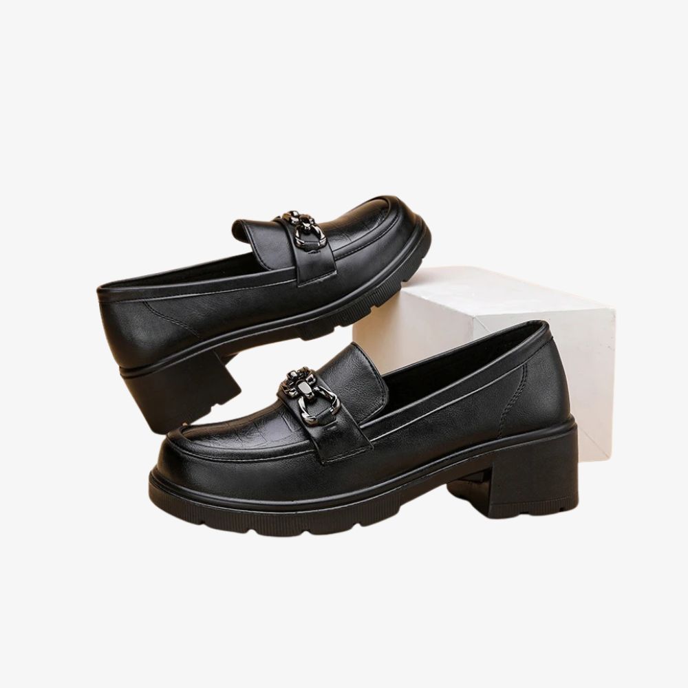 Paar zwarte loafers met hak, waarvan een schoen op een wit blok staat.
