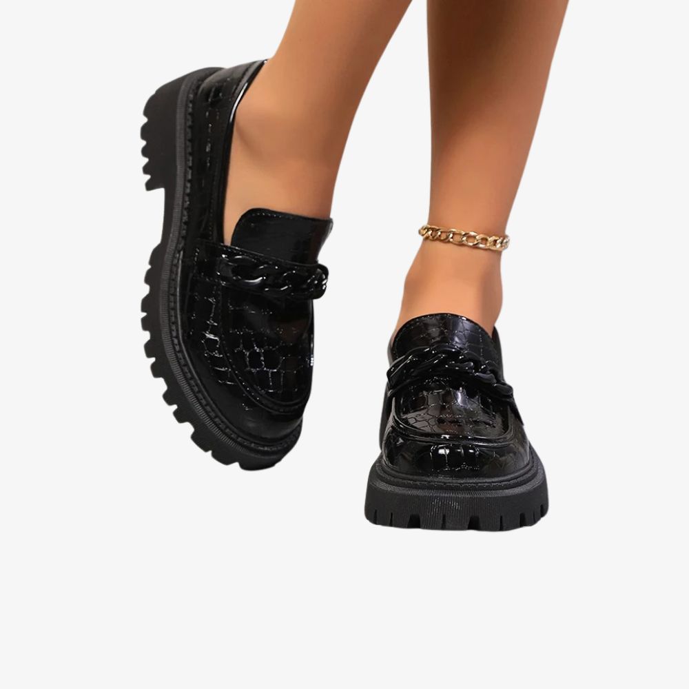 Paar zwarte gelakte loafers met krokodillenprint, gedragen door een vrouw waarvan alleen de enkels zichtbaar zijn. Ze draagt een enkelbandje.