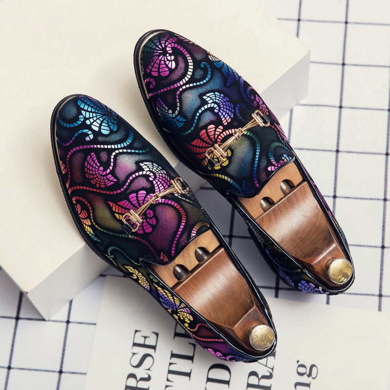 Een paar avondmocassins met veelkleurige arabesken en zilveren stukjes staat op een geruit oppervlak met zwarte en witte letters. Er zitten houten schoenspanners met een gouden knoop aan de binnenkant van de schoenen.