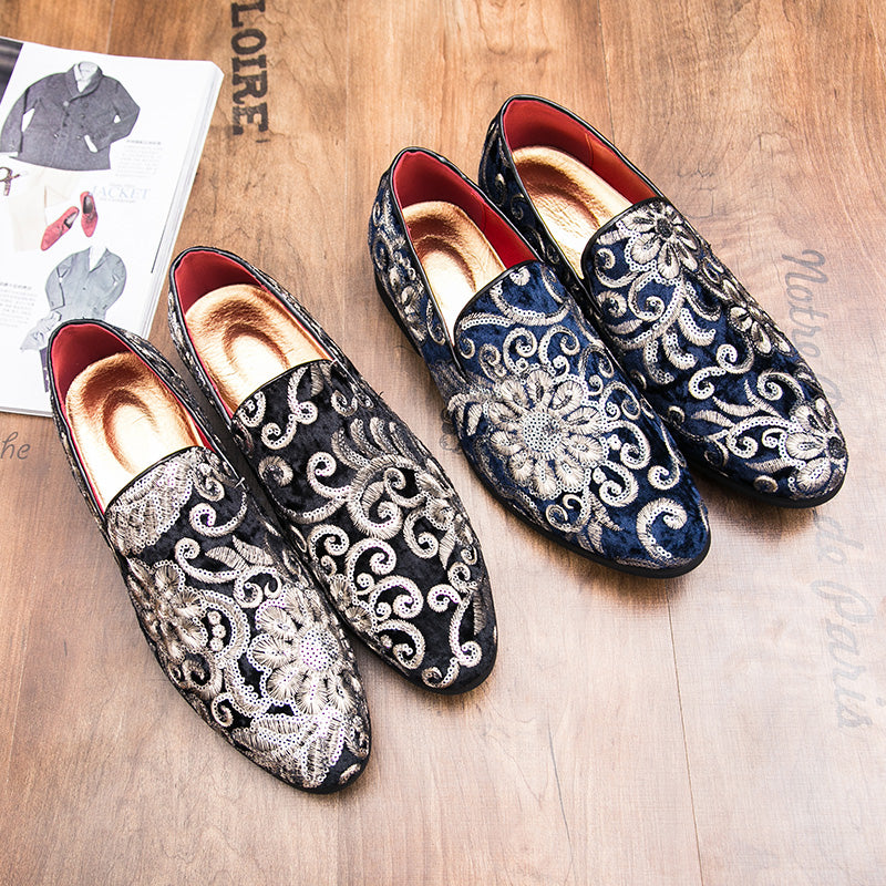 Twee paar zwarte en blauwe mocassins van glanzend fluweel met bloemenborduursel en pailletten en een gouden binnenzool. Ze staan uitgestald op een bruin houten oppervlak naast een tijdschrift over mannenmode