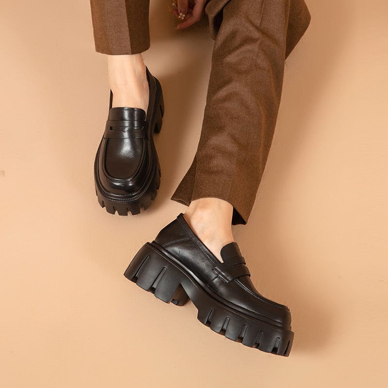 Foto van een paar zwarte loafers gedragen door een vrouw met bruine broek op een roze achtergrond