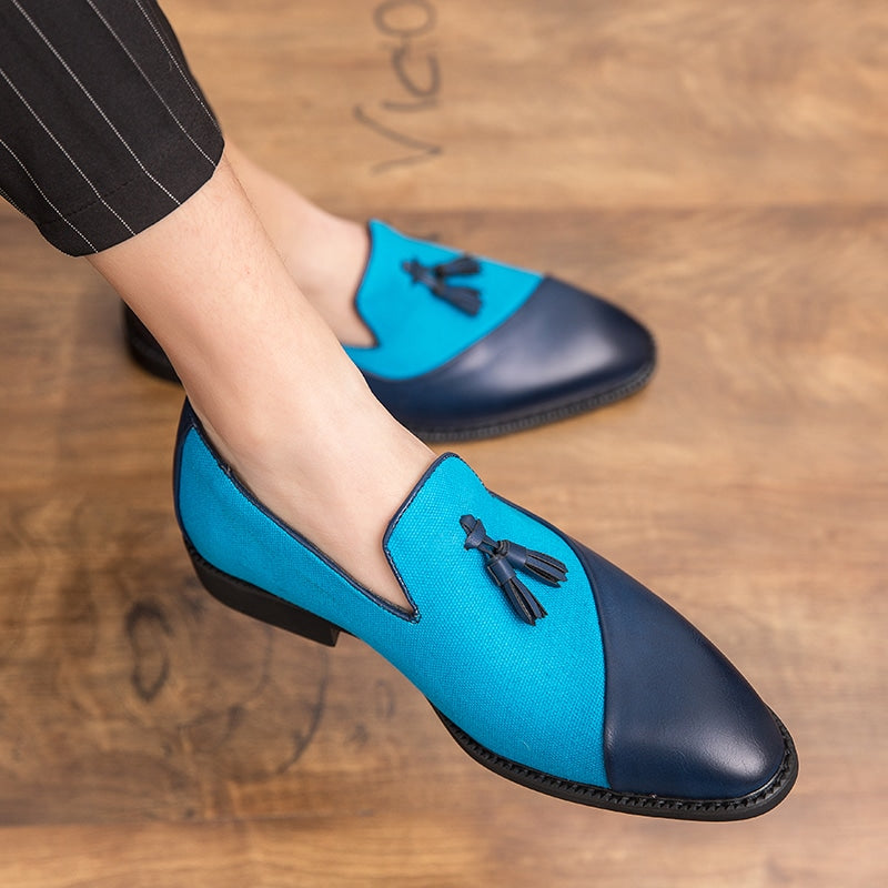 Donker- en lichtblauwe loafers van leer en textiel, contrasterend met decoratieve kwastjes, aan de voeten van een man in kleermakerszit die een zwarte pantalon met fijne witte strepen draagt.