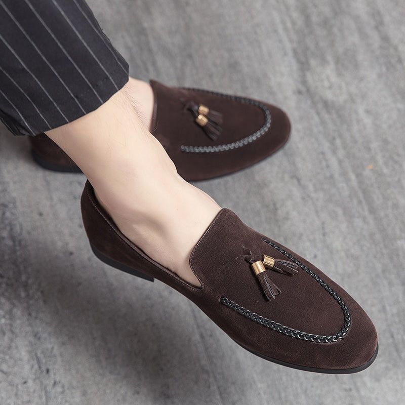 Suède loafers met decoratieve kwastjes en een leren twist op de naad, aan de voeten van een man in kleermakerszit die een zwarte pantalon met dunne witte strepen draagt.