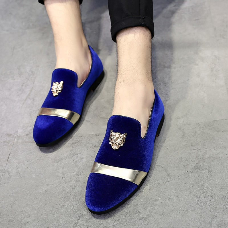 Een paar glanzend blauw fluwelen loafers met een gouden riempje aan de voorkant en een tijgerkop in reliëf op de bovenkant. Gedragen met een zwarte broek op een grijze vloer