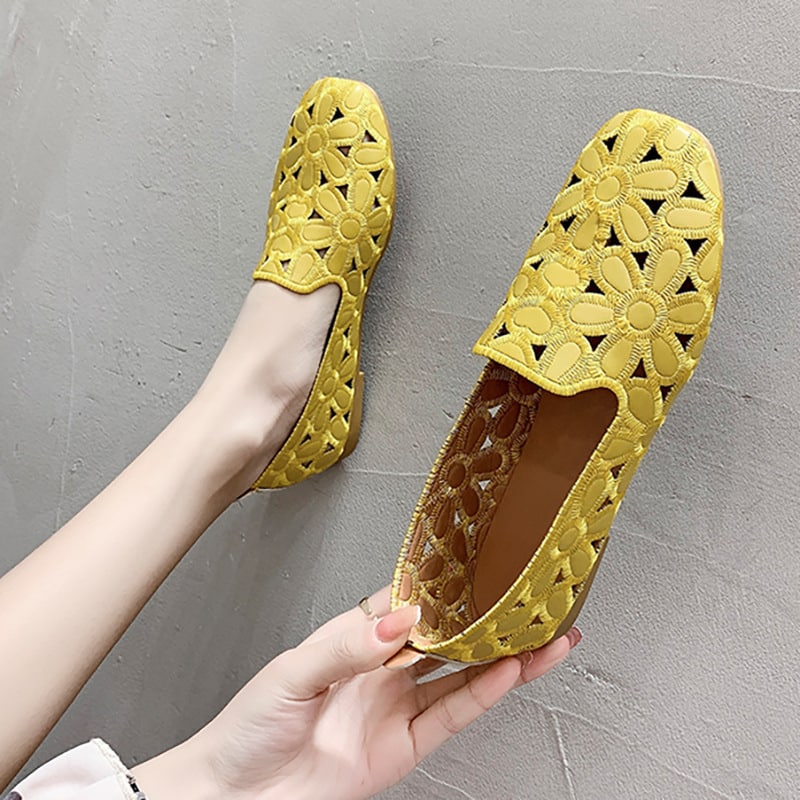 Paar gele mocassins met bloemmotieven. Eén schoen zit in een voet die tegen een grijze muur rust en de andere wordt in een hand gehouden