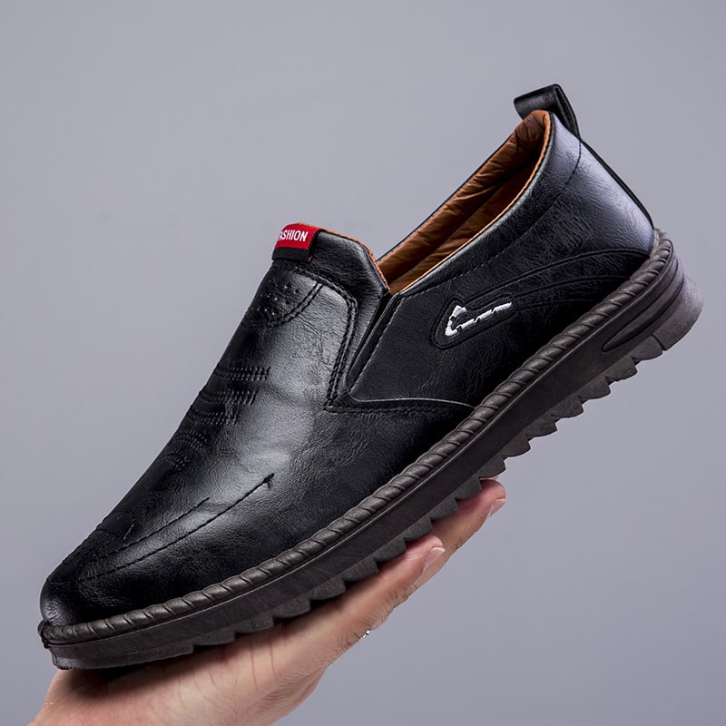 Zwarte loafer met dikke zool, vastgehouden in één hand. Grijze voet.