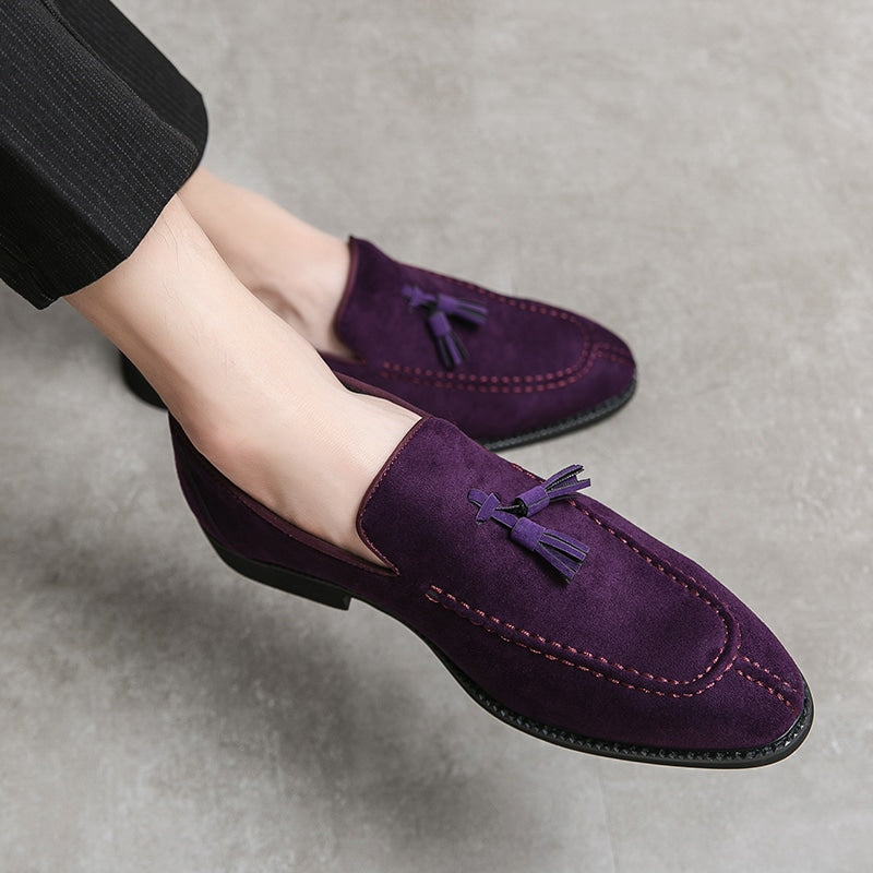Paarse suède loafers met decoratieve kwastjes aan de voeten van een man in kleermakerszit die een zwarte broek met dunne witte strepen draagt.