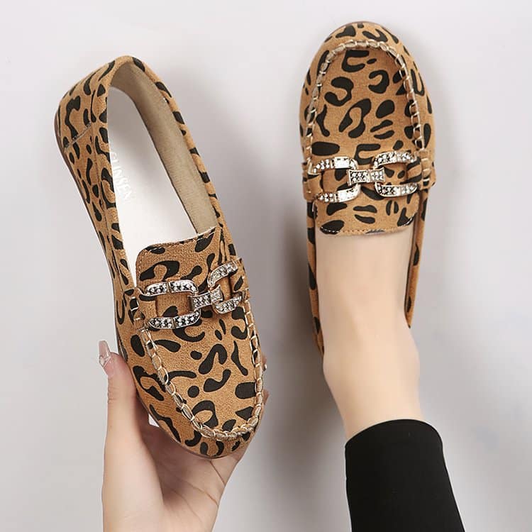 Een paar afgeronde loafers met bruine luipaardprint en zilveren strasringen, één schoen aan de voet met een zwarte panty en de andere in één hand