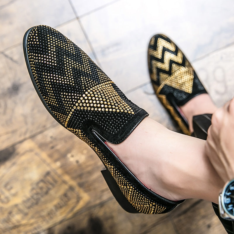 Blootsvoets gedragen, een paar mocassins in zwart met geometrisch borduurwerk van gouden strass. Eén voet rust op een bruine parketvloer.