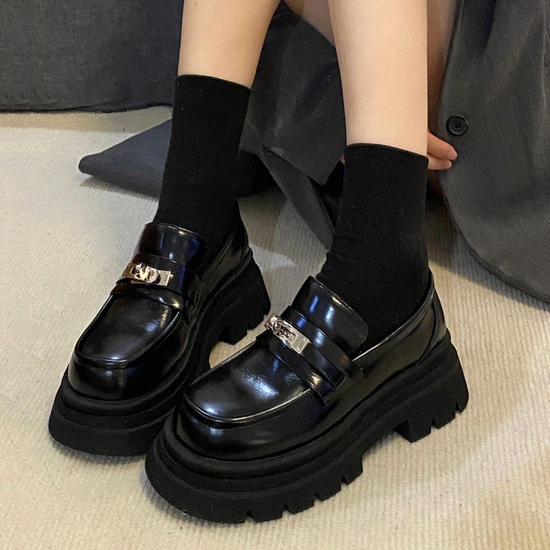 Zwarte mocassins van glanzend glad leer met een dikke zool aan de voeten van een vrouw die op de grond zit en korte zwarte sokken draagt.