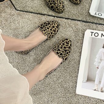 Een paar eenvoudige loafers met luipaardprint van bovenaf gezien, gedragen met een witte rok en blote voeten. De vloer is beige tapijt en er ligt een wit tijdschrift naast
