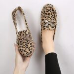Paar mocassins in luipaardprint met stiksels en zilveren strasringen bovenop. Eén schoen in één voet en de andere in één hand.