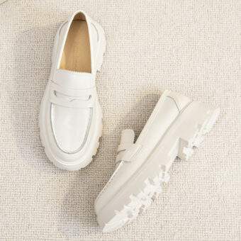 Een paar eenvoudige witte gladleren loafers met dikke witte zolen op een lichtbeige vloer.