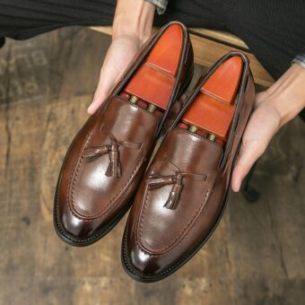 Donkere bruine leren loafers met puntige tenen en decoratieve kwastjes in de handen van een man die ze boven een houten vloer houdt.
