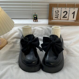 Een paar mocassins van glad zwart leer op een wit bed, met een grote strik bovenaan en beige sokjes die de schoen opvullen.