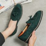 Smaragdgroene suède loafers met decoratieve kwastjes aan de voet in de hand van iemand die op een lichtgrijze vloer zit.