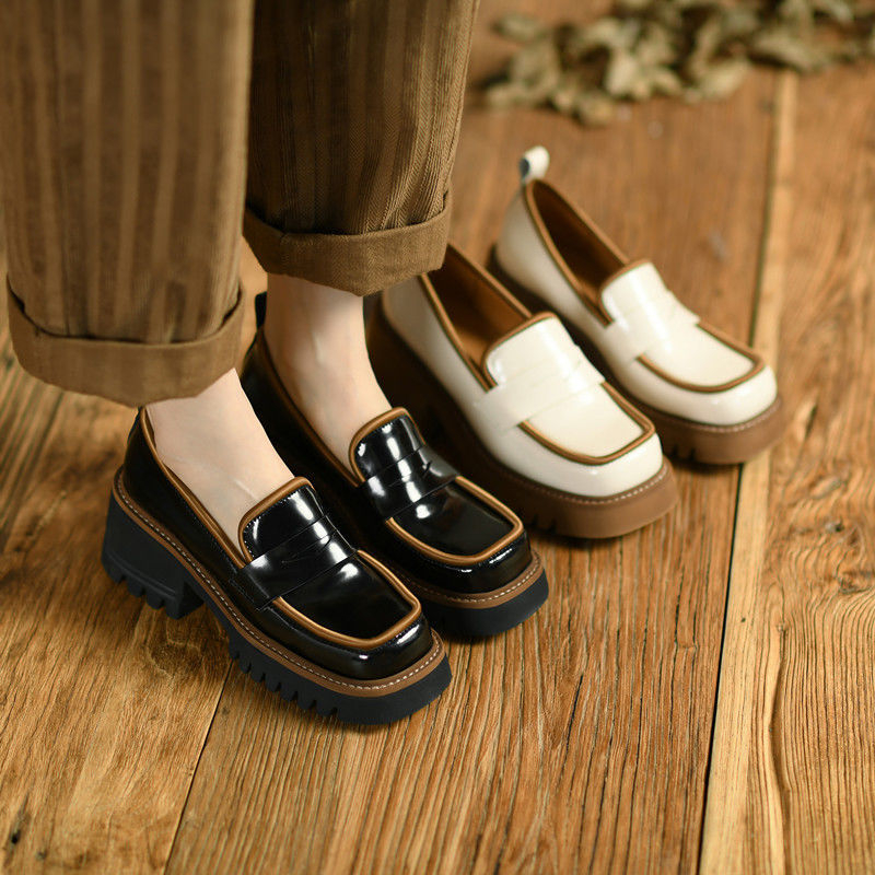 Twee paar contrasterende lakleren loafers, de ene beige en bruin, de andere zwart en bruin, aan de voeten van een vrouw die op een parketvloer staat en een wijde bruine gestreepte broek draagt.