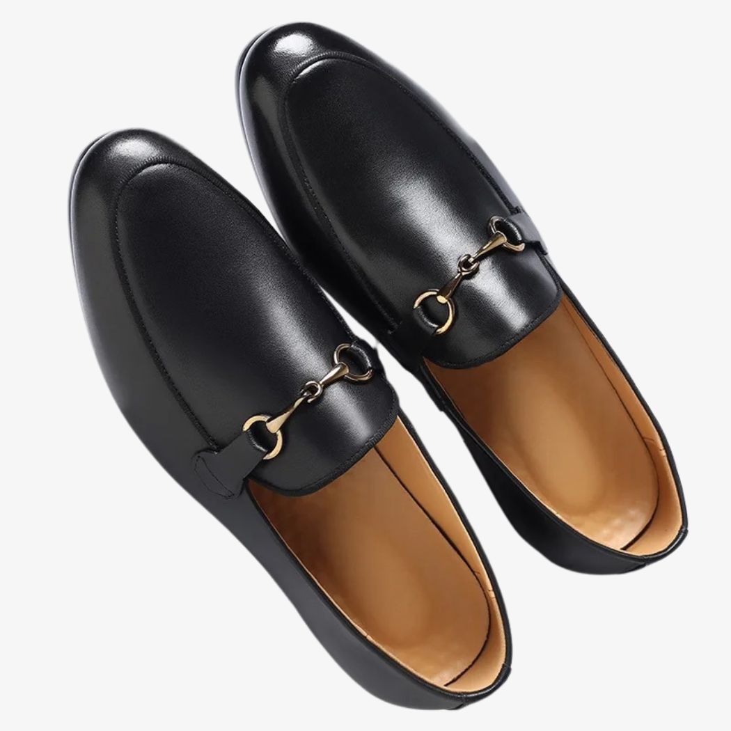 Zwarte mocassin met gouden gesp van veganistisch leer op een gebroken witte achtergrond.